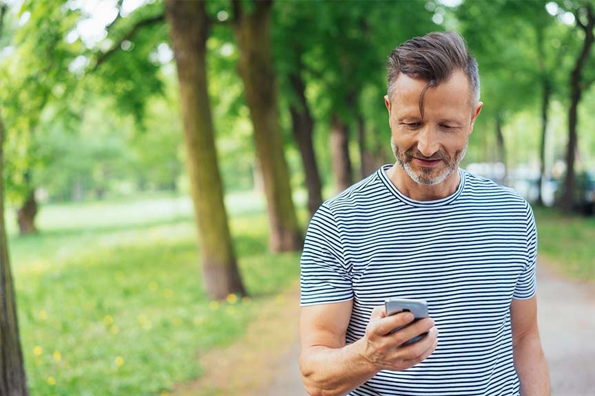 A man, walking between trees, looking at his phone.